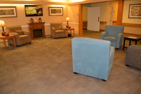 A cozy common area with several armchairs arranged around a small electric fireplace. The room has warm lighting from table lamps, framed artwork on the walls, and a carpeted floor. There is a doorway leading to another room in the background.