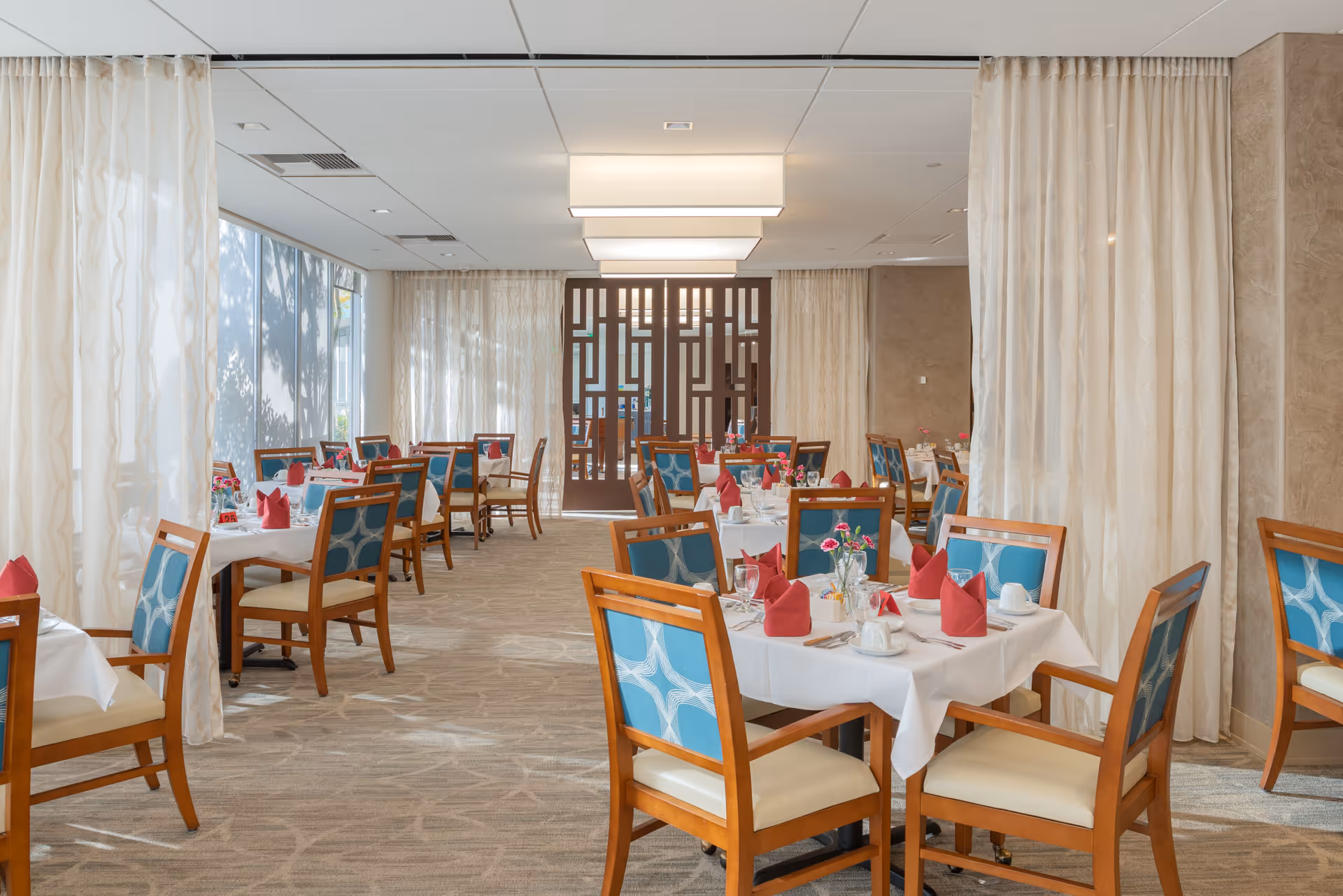 A bright and elegant dining room with multiple tables set with white tablecloths, red folded napkins, glassware, and small flower arrangements. The chairs have wooden frames with blue patterned upholstery. Large windows with sheer curtains allow natural light to fill the room, and modern rectangular ceiling lights provide additional illumination.