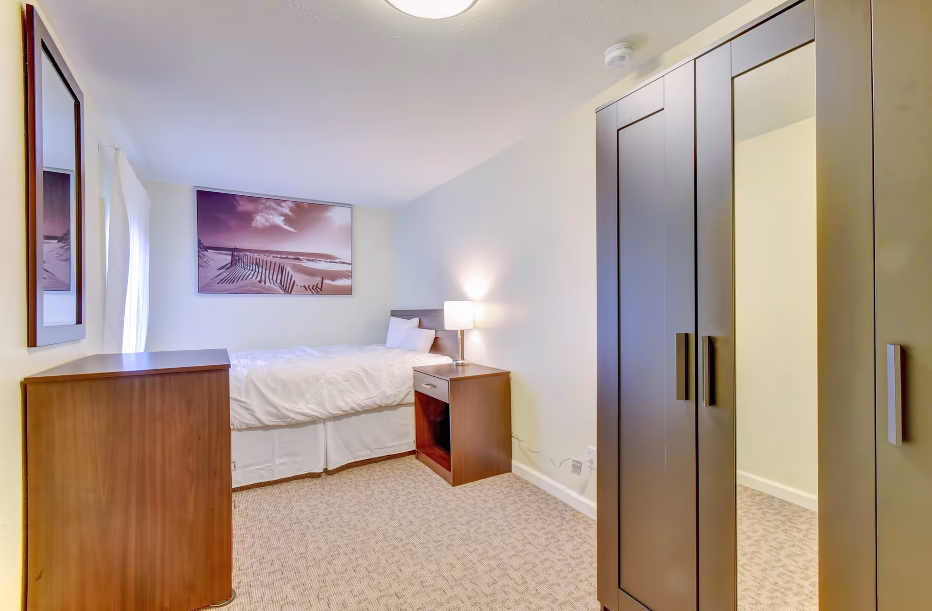 A small, neatly arranged bedroom with a single bed against the far wall, covered with white bedding. Next to the bed is a wooden nightstand with a lamp on it. On the right side of the room, there is a tall wardrobe with mirrored doors. On the left side, there is a wooden dresser and a framed picture hanging on the wall. The room is carpeted and has light-colored walls.