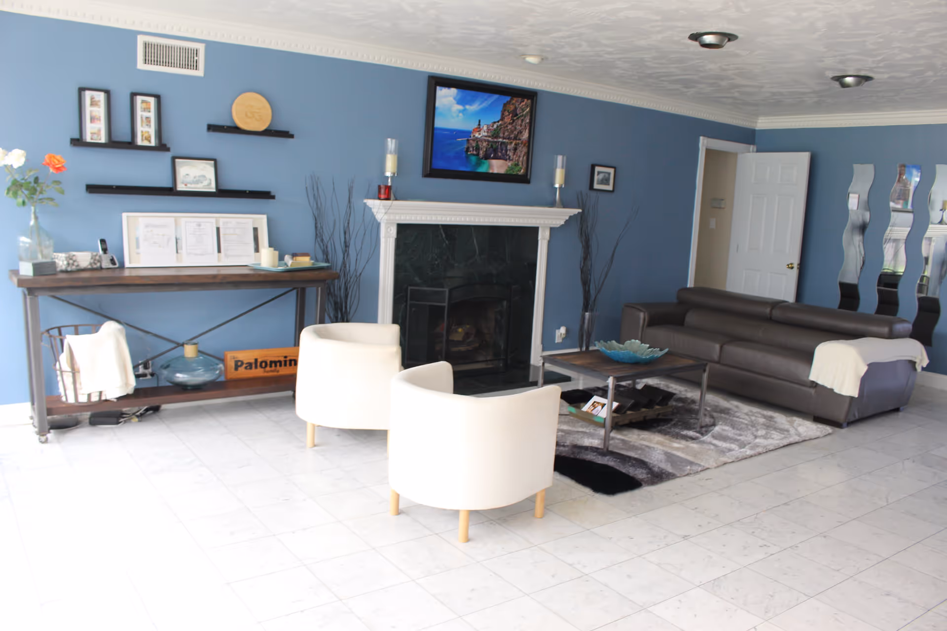 A cozy living room with blue walls, a white tiled floor, and a white ceiling. The room features a dark brown leather sofa with a white throw blanket, two white armchairs, a wooden coffee table with a decorative bowl, and a fireplace with a black marble surround and white mantel. Above the fireplace is a framed picture of a coastal scene. On the left side, there is a wooden console table with framed pictures, candles, and a vase with flowers. Decorative wall shelves and tall vases with branches flank the fireplace. A set of wavy mirrors is mounted on the wall near a white door.