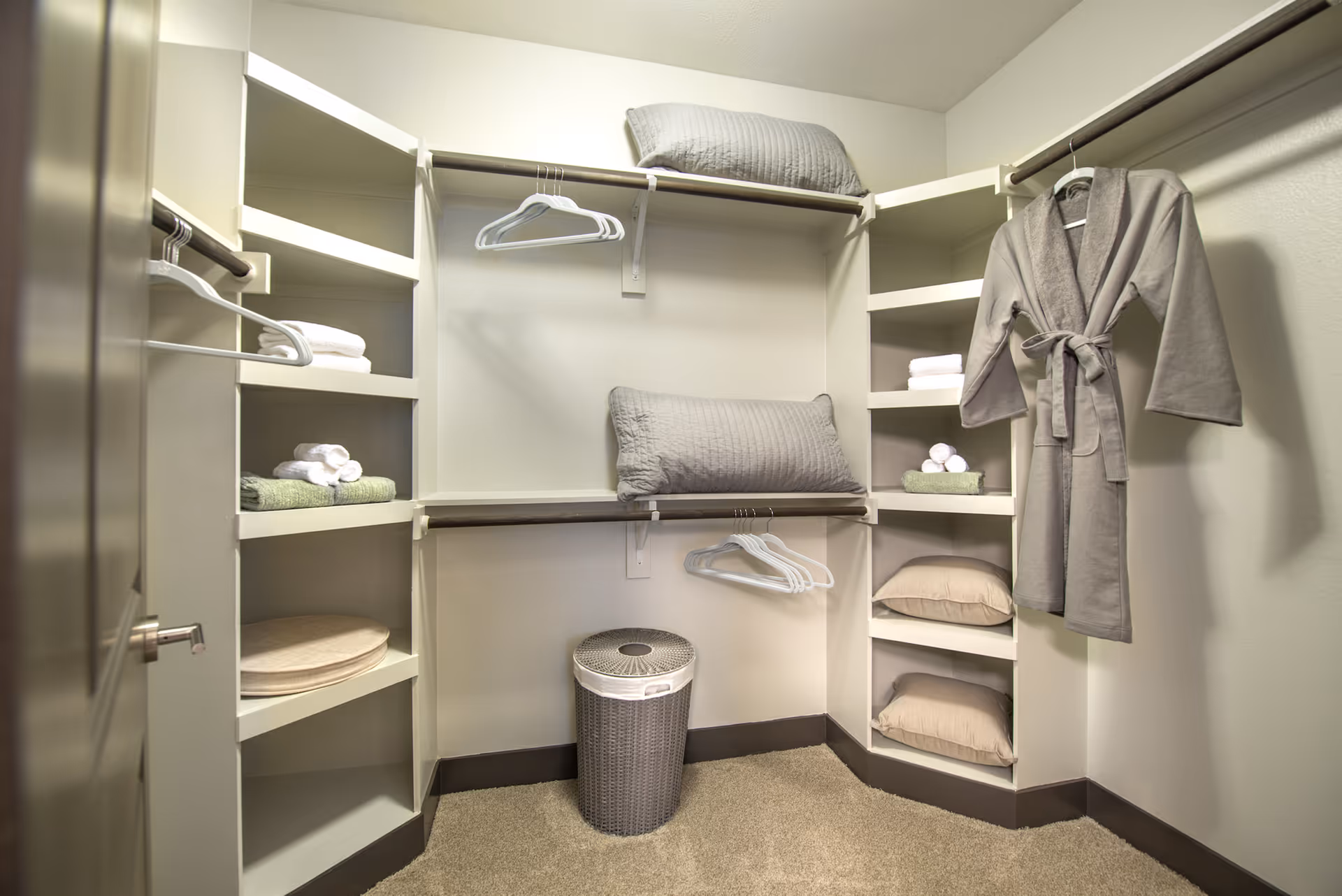 A walk-in closet with white shelves and hanging rods. The shelves hold folded towels, pillows, and a round cushion. A gray bathrobe hangs on the right side. There are several empty white hangers on the rods and a gray laundry basket on the carpeted floor.