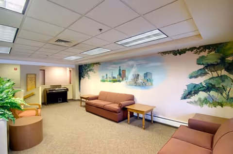 A senior living facility common area with two brown sofas and a wooden side table between them. The walls feature a mural of a city skyline and a fountain, along with painted greenery. There is a piano against the far wall and a potted plant in the foreground. The ceiling has fluorescent lighting panels.