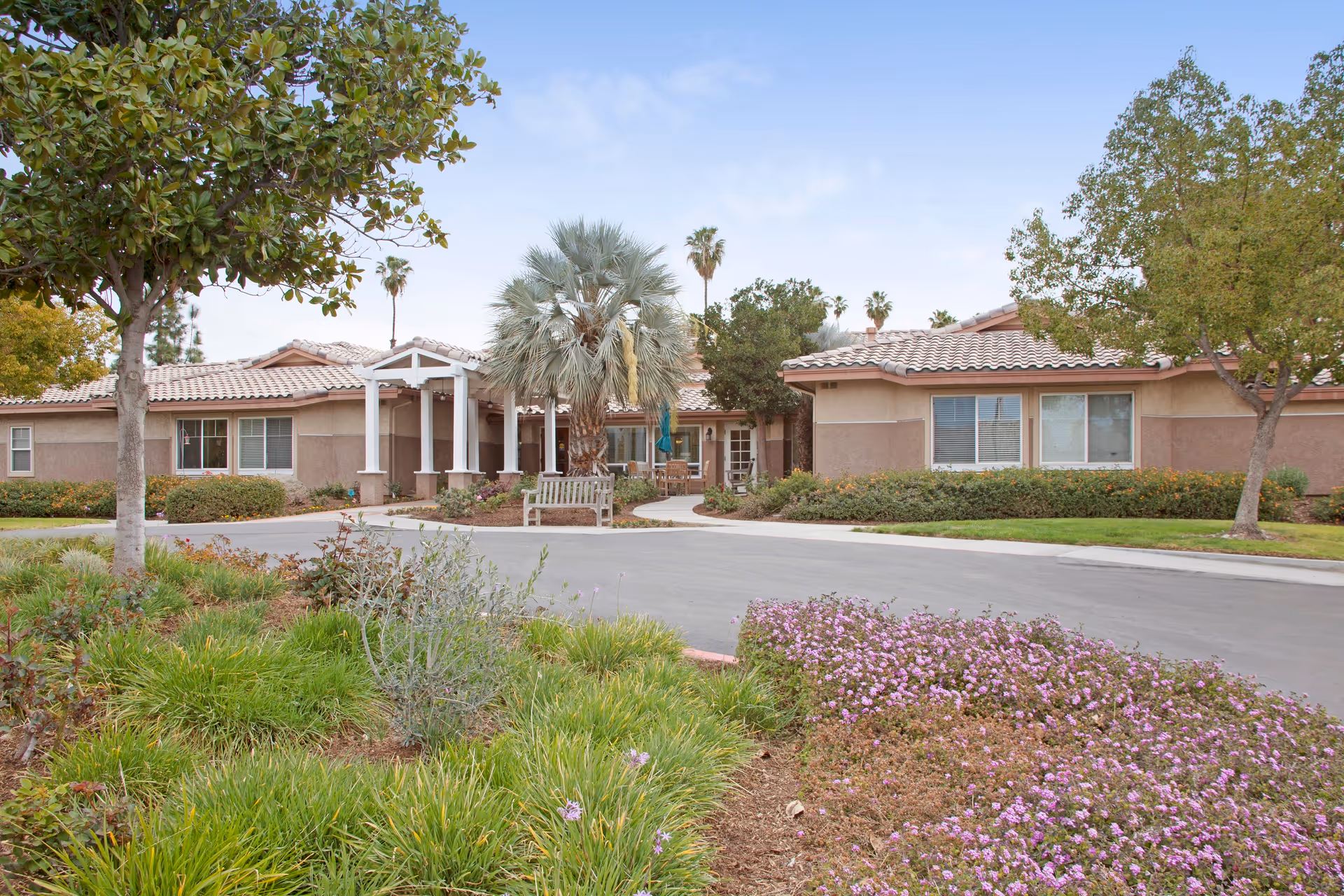 Single-story senior living building with a tiled roof, central covered entrance and landscaped grounds with trees and flowering plants.