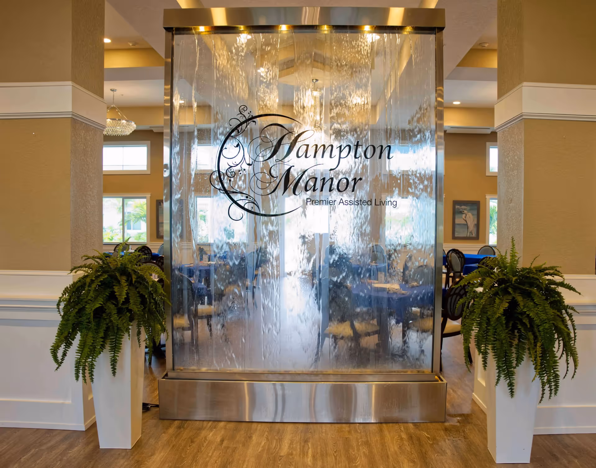 Indoor water feature with flowing water and the text 'Hampton Manor Premier Assisted Living' displayed on the glass panel, flanked by two large potted ferns, with a dining area visible in the background.