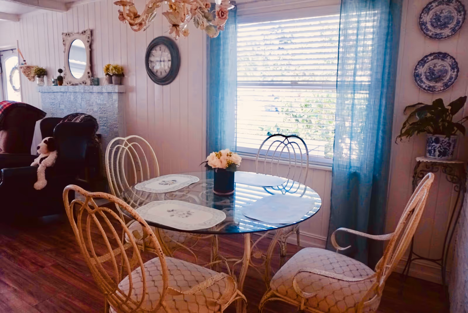 Airy dining nook featuring a round glass table with four wicker chairs, teal curtains over a large window, and decorative plates and furnishings.