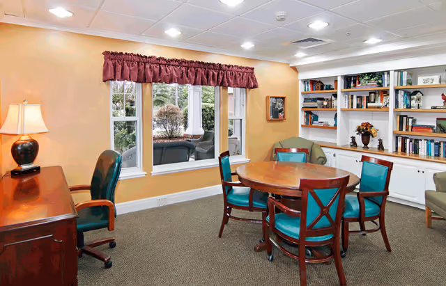Cozy communal sitting area with a round wooden table and teal chairs, bookshelves, a desk lamp, and a window looking out to greenery.