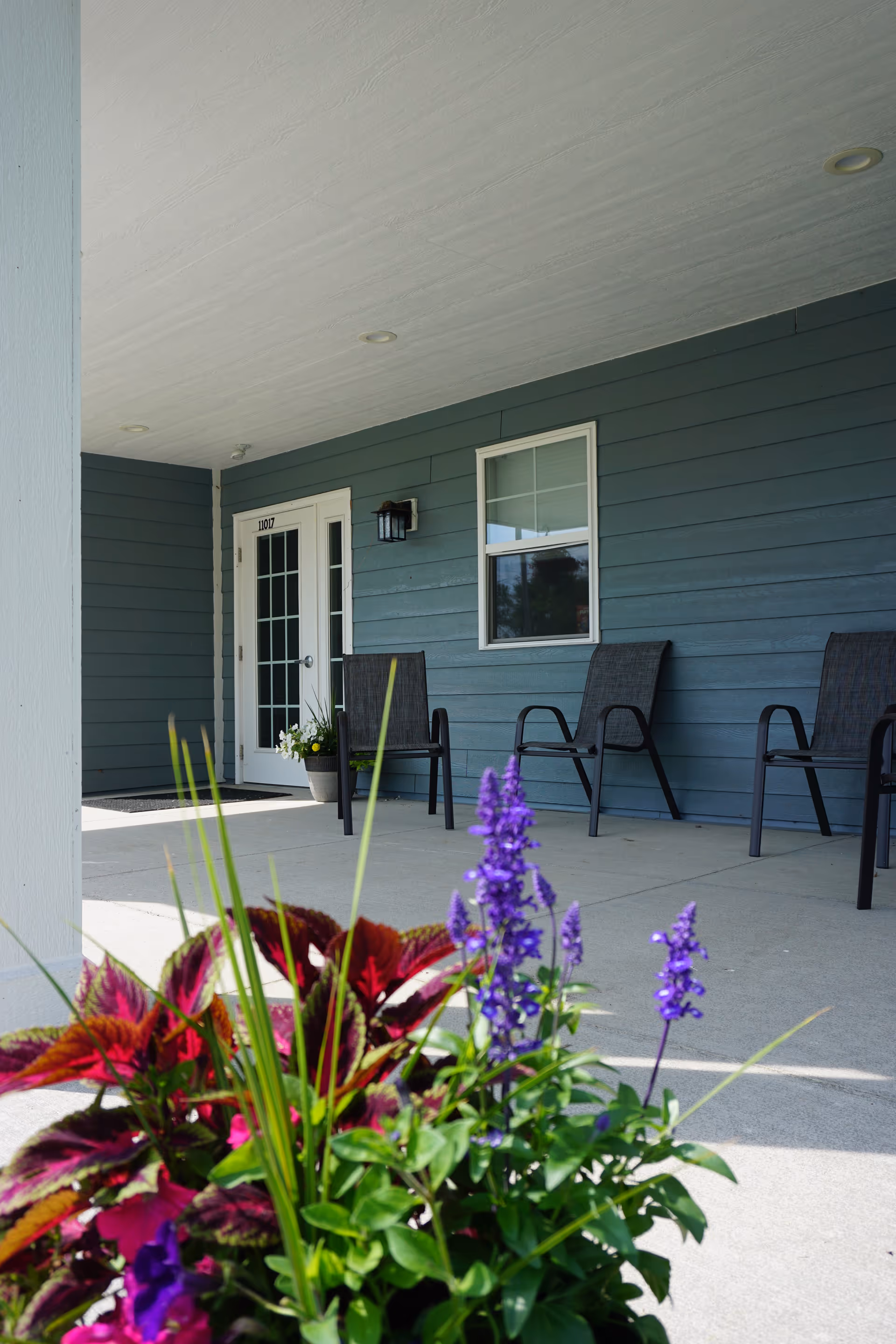 Covered front porch with chairs, an entrance door and window, and potted purple flowers in the foreground.