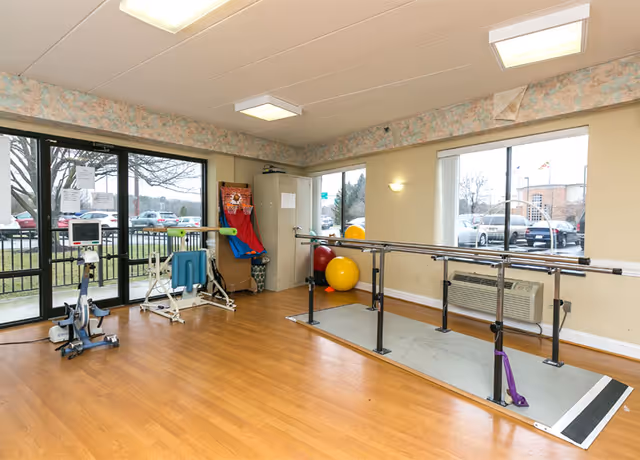A rehabilitation exercise room with wooden flooring, parallel bars for walking practice, exercise balls, a stationary bike, and other physical therapy equipment. Large windows provide natural light and a view of parked cars outside.
