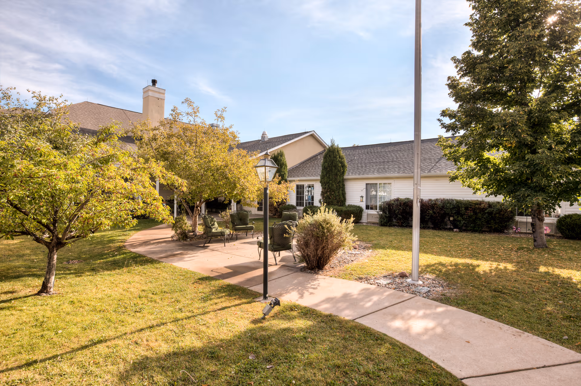 Outdoor courtyard area at Brookdale Broadmoor with a paved walkway, green lawn, trees, shrubs, and several green cushioned chairs arranged around a small table under a tree. The building exterior is visible in the background under a partly cloudy sky.