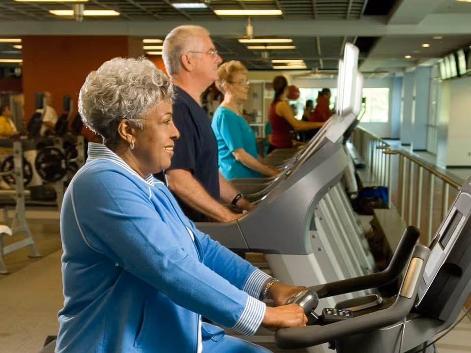 Several elderly individuals exercising on treadmills in a fitness center, with a woman in a blue jacket in the foreground smiling as she uses the equipment.