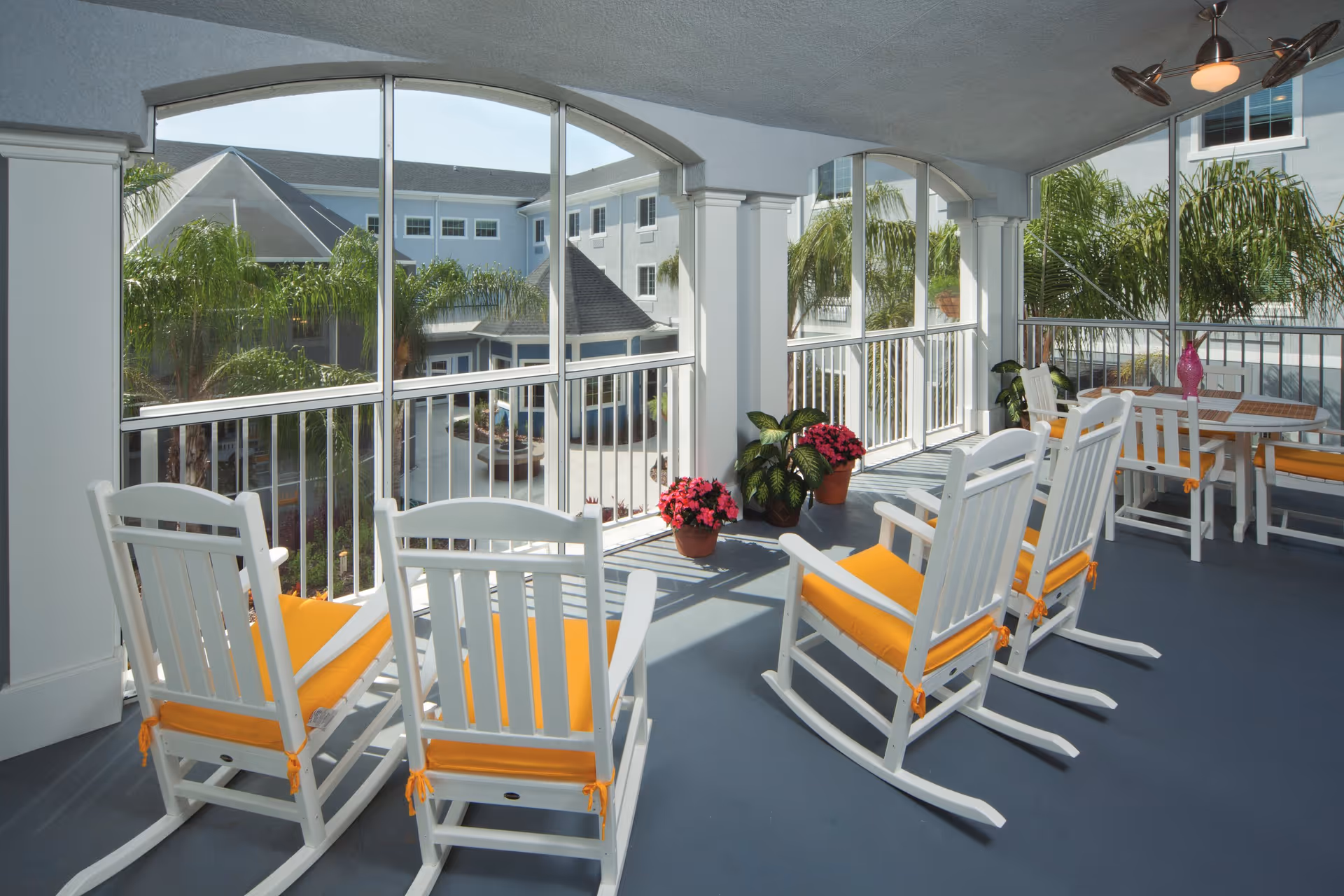 Covered screened balcony with white rocking chairs with orange cushions, potted plants, and a view of the courtyard and surrounding buildings.