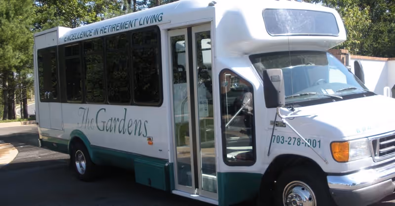 A white and green shuttle bus parked on a driveway with the text 'The Gardens' and 'Excellence in Retirement Living' written on its side. The bus has large windows and a front door for passengers. Trees and a building are visible in the background.
