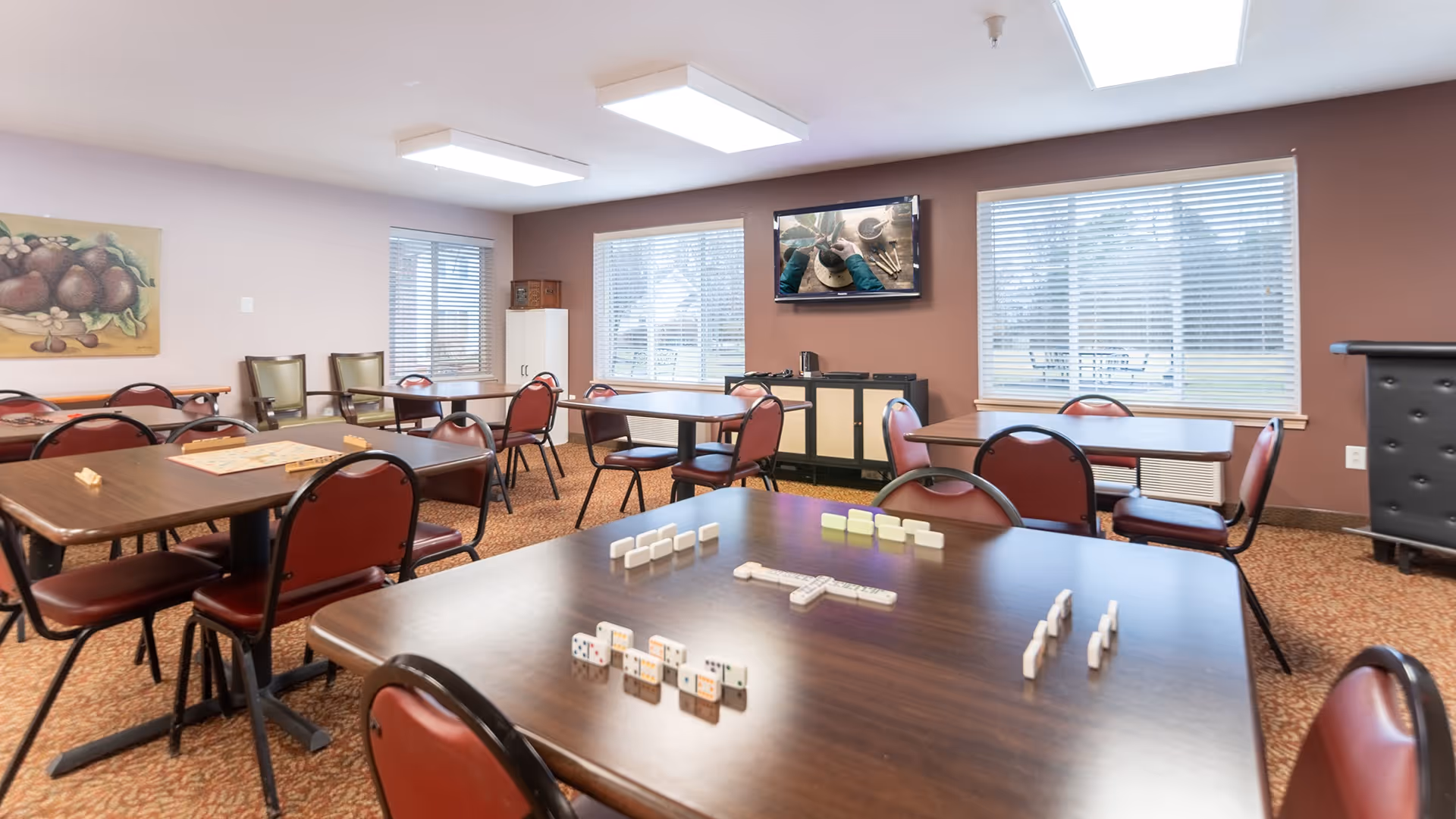 A well-lit common room with multiple tables and chairs arranged for group activities. Dominoes are set up on the closest table, and a television is mounted on a brown accent wall showing a pottery-making video. Large windows with blinds allow natural light to fill the room, and a painting of fruit hangs on one wall.