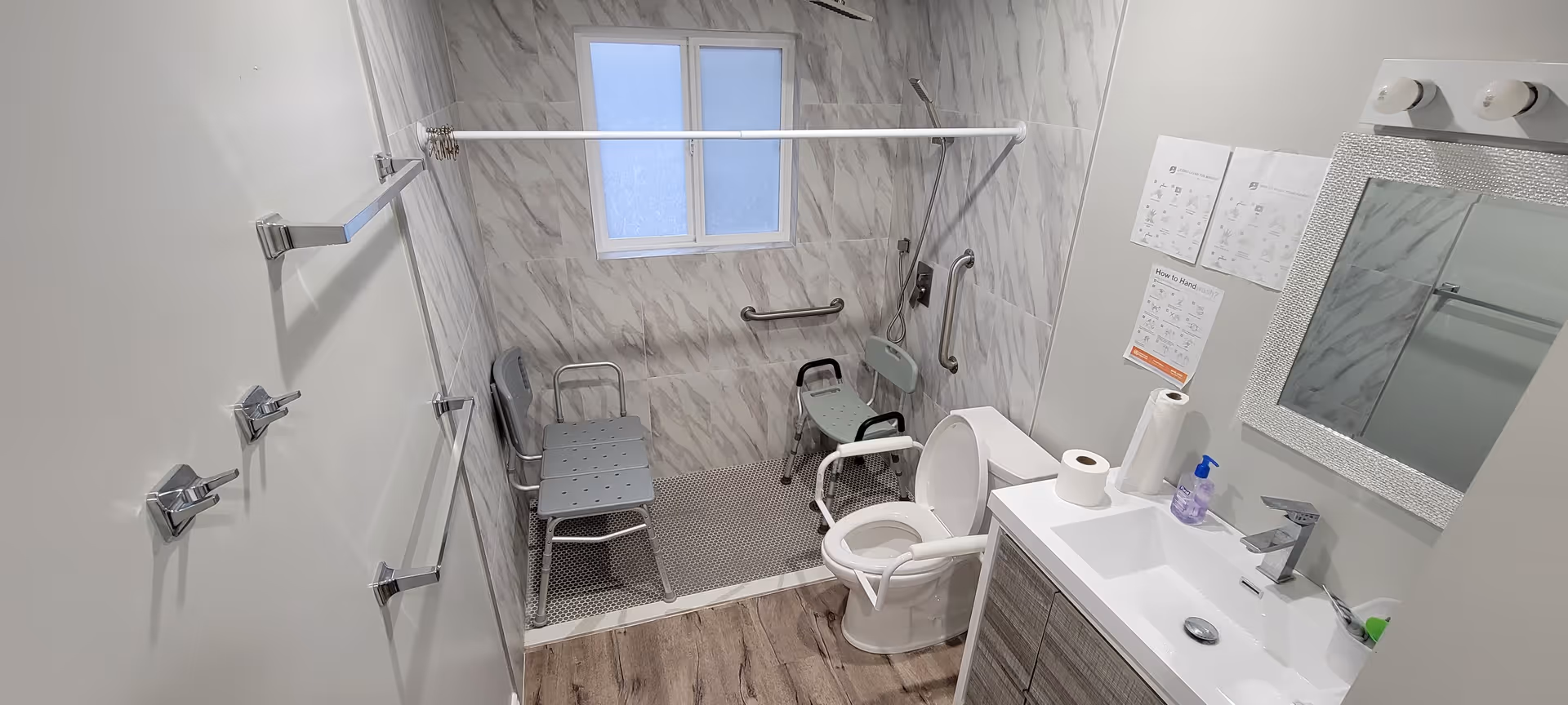 A bathroom designed for accessibility with a walk-in shower area featuring two shower chairs and grab bars. The toilet has arm supports for assistance, and there is a sink with a mirror above it. Instructions on hand washing are posted on the wall next to the mirror. The bathroom has a window and a wood-look floor.