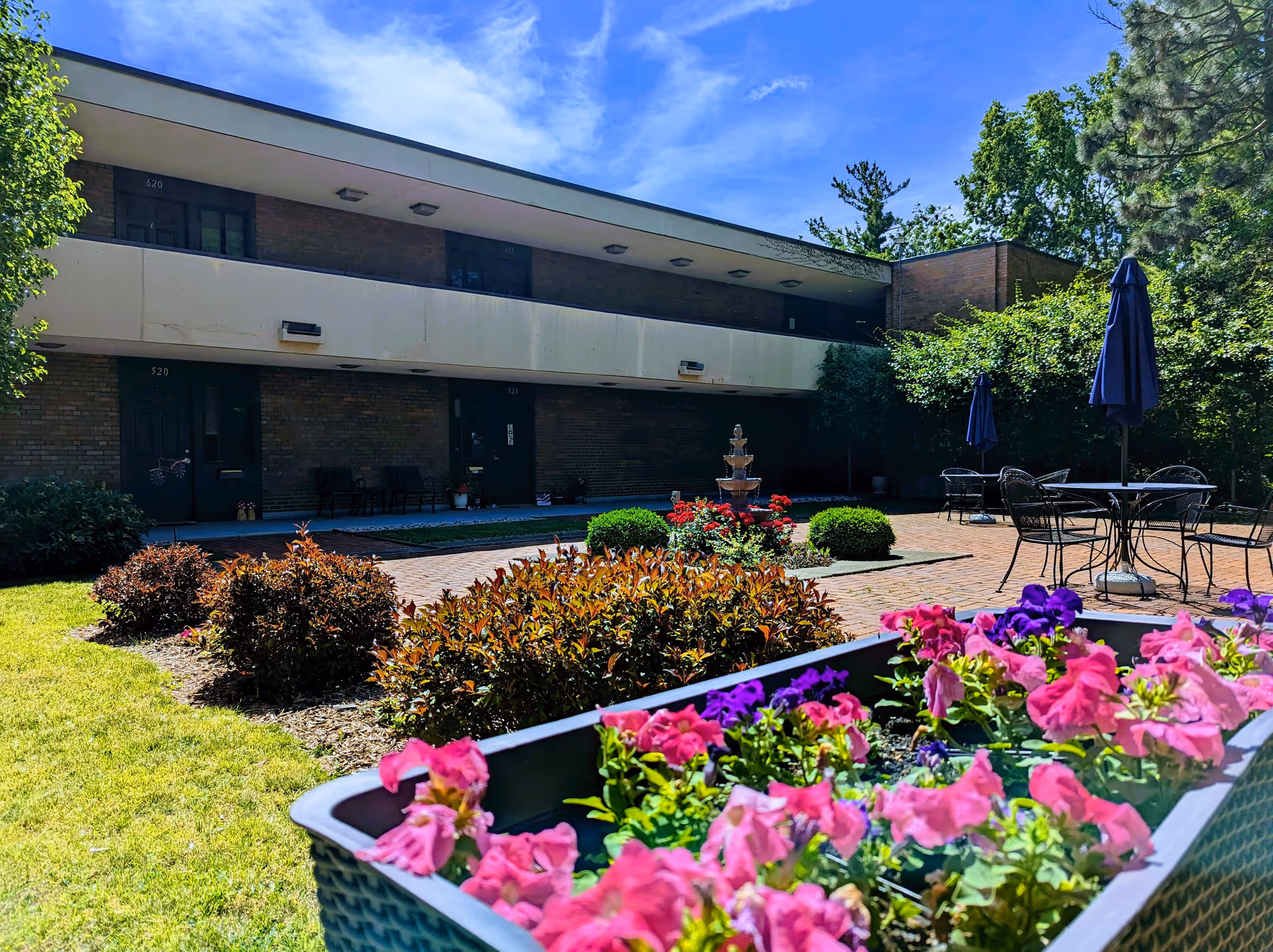 Outdoor courtyard area of Plum Landing Retirement Community featuring a flower planter with pink and purple flowers in the foreground, neatly trimmed bushes, a brick-paved patio with a multi-tiered fountain, and several tables with chairs and closed umbrellas. The two-story building with dark doors and windows is visible in the background under a blue sky with some clouds.