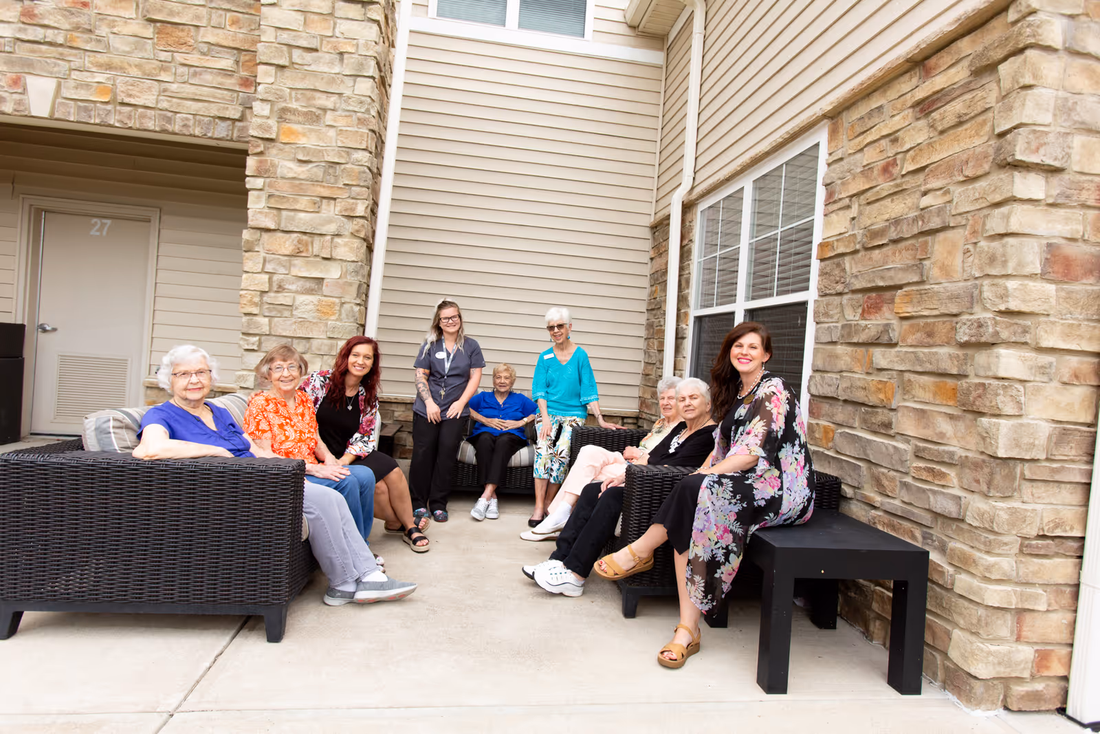 A group of elderly women and a caregiver sitting and standing together on outdoor patio furniture against a stone and siding wall, smiling and enjoying each other's company.