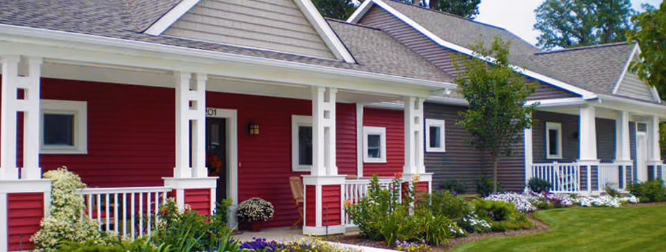Row of single-story residential buildings with front porches, white railings, and well-maintained gardens with various plants and flowers in front. The buildings have different colored sidings including red and gray, with a green lawn in the foreground.