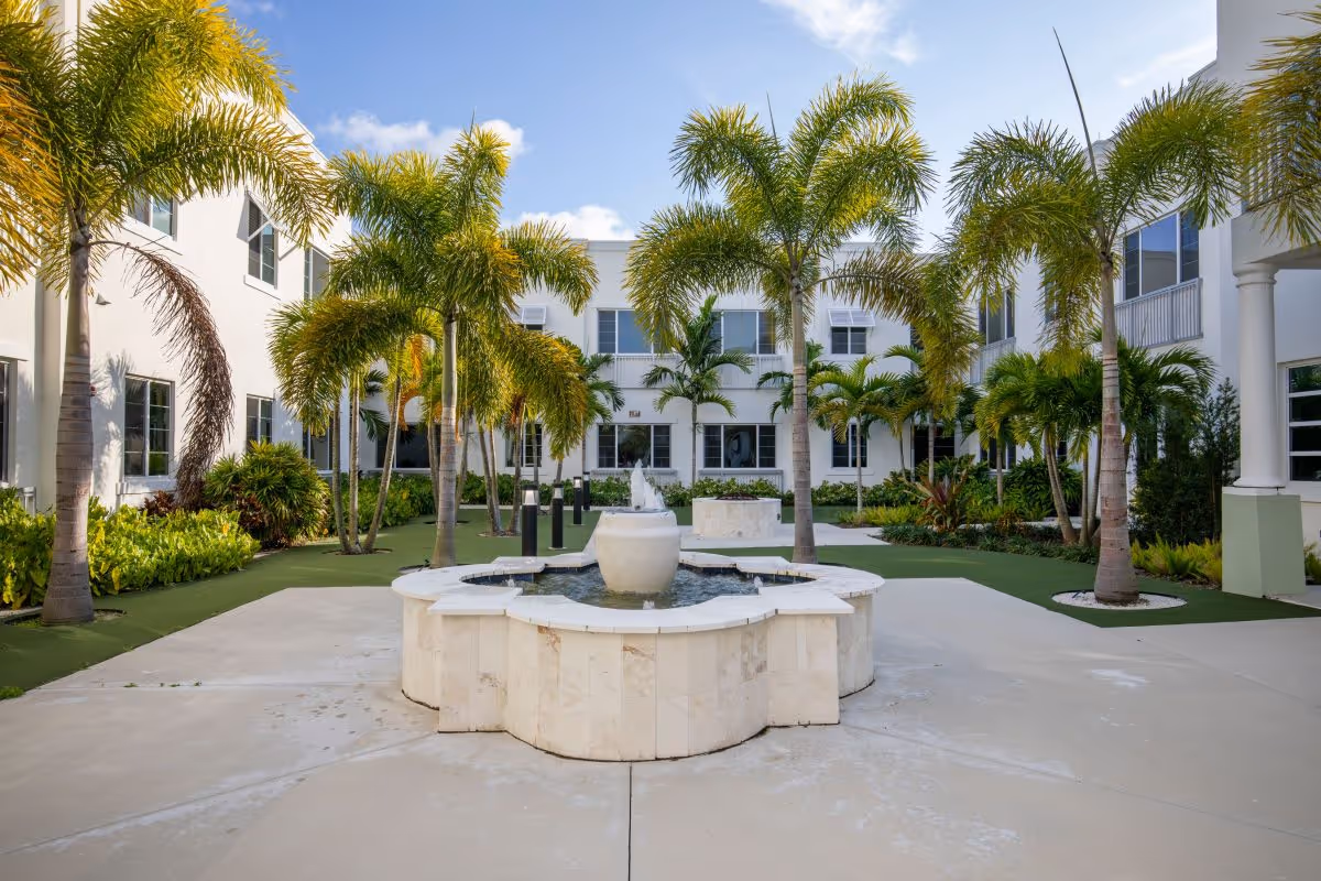 Courtyard with a central stone fountain surrounded by palm trees and a two-story white building under a blue sky.