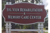 Outdoor sign for Syl-View Rehabilitation & Memory Care Center mounted on white posts with trees and greenery in the background.