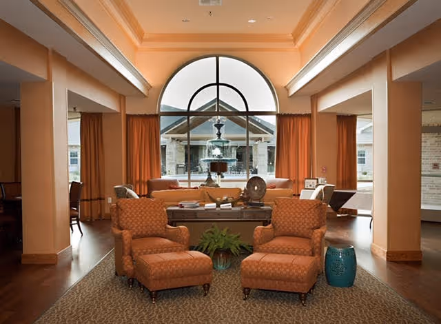 A cozy living room area with two patterned armchairs and a small blue side table in the foreground. Behind the chairs is a coffee table with books and decorative items, and a large window with an arched top showing an outdoor fountain and building exterior. The room features warm tones with orange curtains and walls, and wooden flooring.