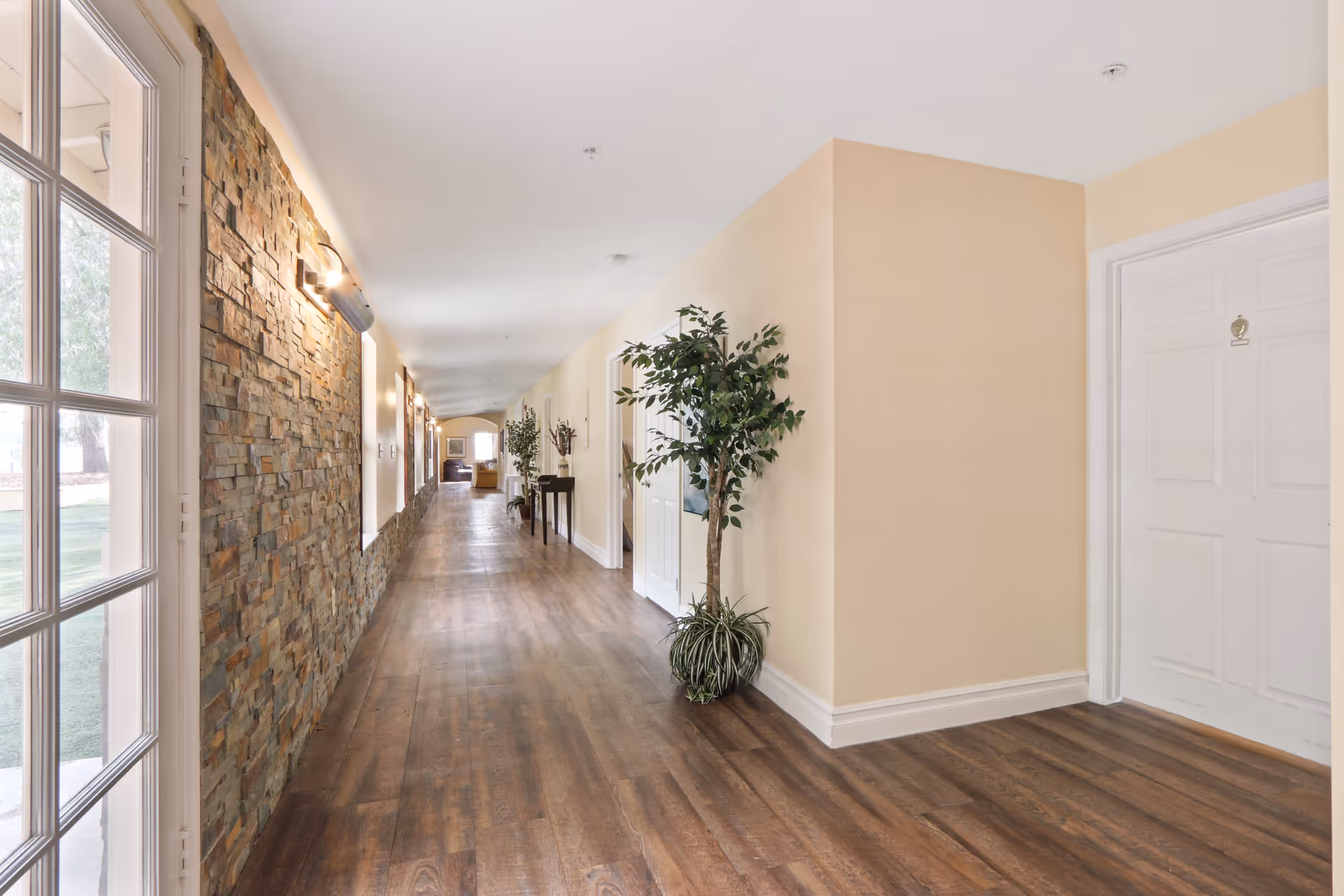 A long, well-lit hallway in a senior living facility with wooden flooring, beige walls, and a stone accent wall on the left. There are several white doors along the right side, potted plants, and wall-mounted lights. At the far end, a seating area with chairs is visible.