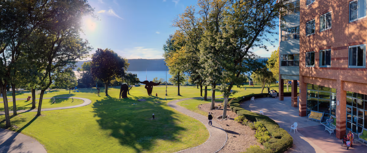 Outdoor view of a senior living facility with a well-maintained green lawn, walking paths, trees, and sculptures. The building is visible on the right side with large windows and a patio area with chairs and benches. The sun is shining brightly in a clear blue sky.