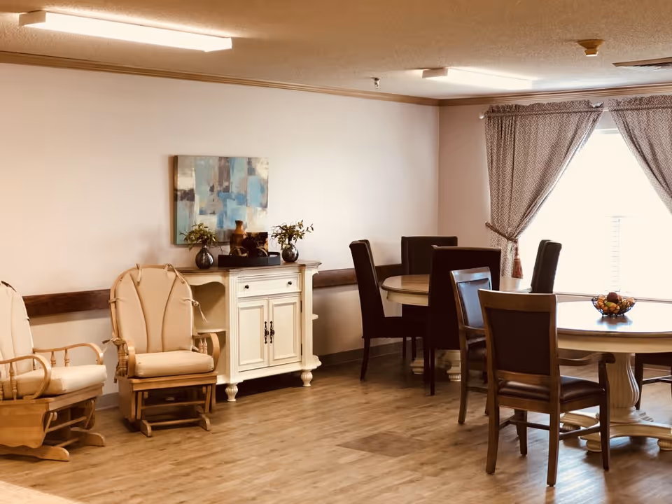 A cozy interior room with wooden flooring featuring two cushioned rocking chairs on the left, a white cabinet with decorative items and a painting above it on the wall, and two round dining tables with dark cushioned chairs near a window with patterned curtains.