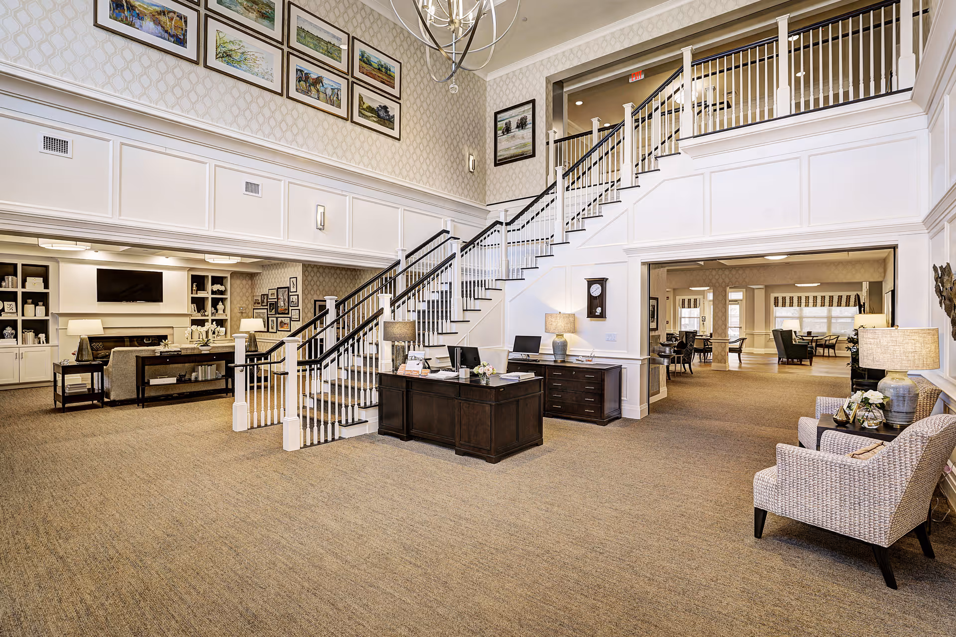 Spacious and well-lit senior living facility interior featuring a central staircase with white railings and dark handrails. The room has beige carpeting, patterned wallpaper, and multiple framed paintings on the walls. There is a reception desk with lamps and computers, a seating area with armchairs and side tables, and an open view into a dining area with tables and chairs. The overall decor is elegant and welcoming.