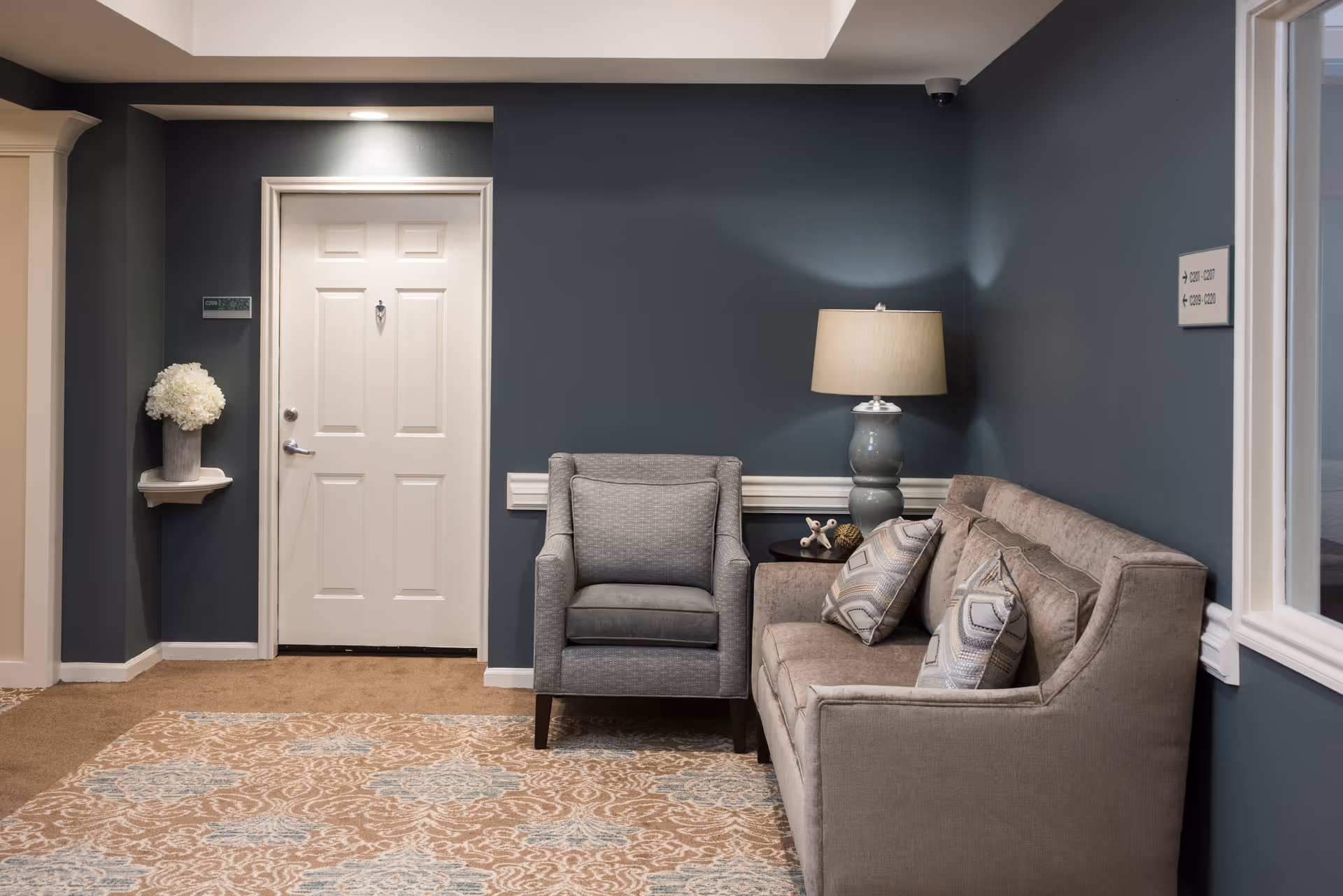 A cozy seating area in a senior living facility with a gray armchair and a beige sofa adorned with patterned cushions. A table lamp with a beige shade sits on a side table between the chair and sofa. The walls are painted dark teal, and a white door is visible in the background next to a small shelf holding a vase of white flowers. A patterned area rug covers the floor.