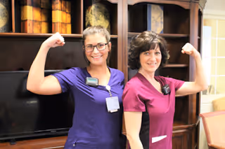 Two women wearing scrubs, one in purple and one in maroon, standing indoors in front of a wooden cabinet and television, smiling and flexing their arm muscles in a playful, confident pose.