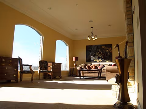 A spacious living room with large arched windows letting in natural light. The room features a beige carpet, a sofa with multiple cushions, a coffee table, two wooden chairs, a chest of drawers, a side table with a lamp, a chandelier, and a decorative painting on the wall. There is also a tall decorative vase near a stone wall on the right side.