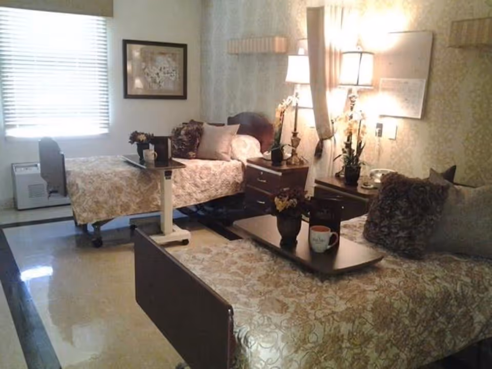 Shared patient bedroom with two beds, nightstands, lamps, and decorative pillows.