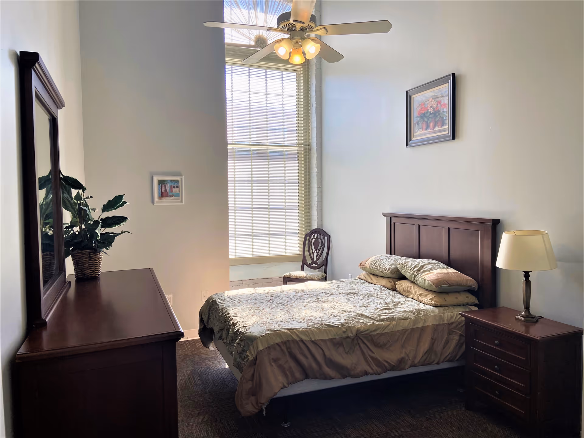Sunlit bedroom with a neatly made bed, wooden headboard, nightstand with lamp, dresser with mirror and potted plant, tall window and ceiling fan.