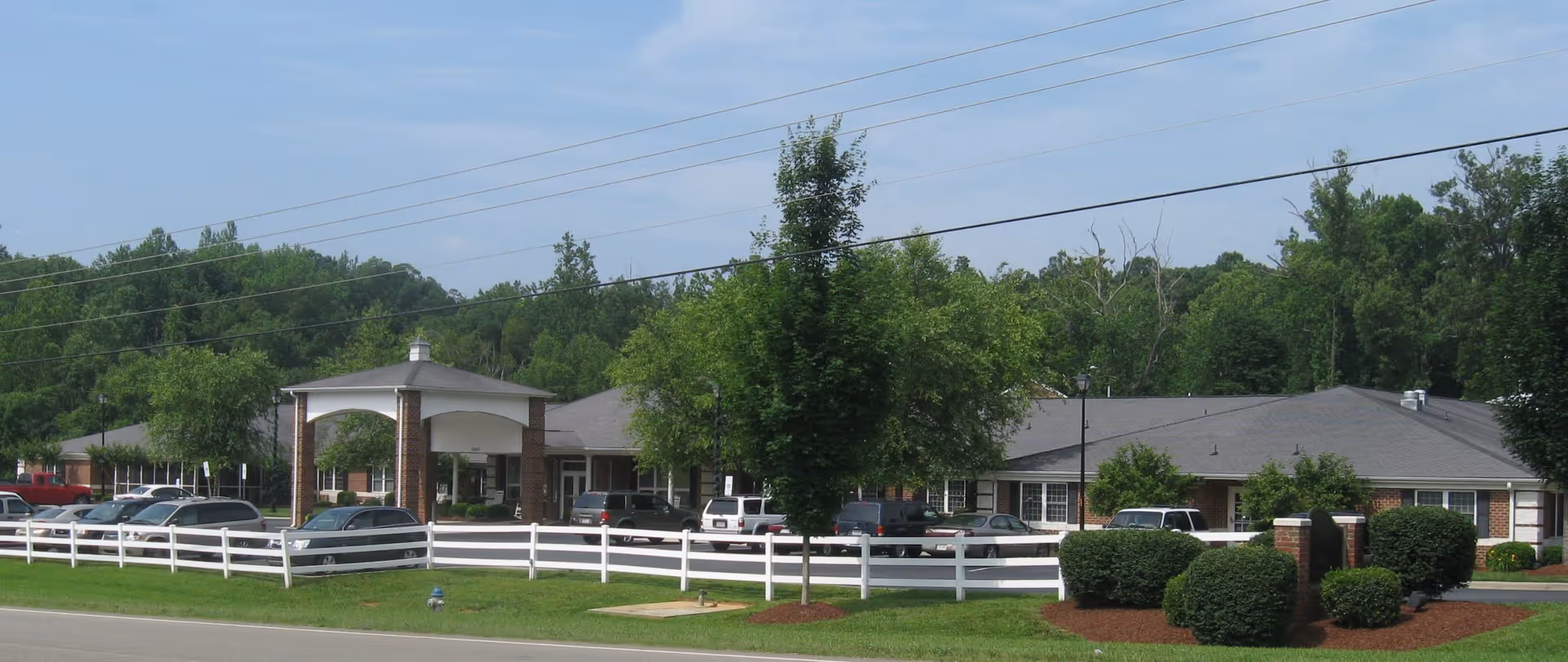 Exterior view of Roxboro Assisted Living facility showing a single-story brick building with a covered entrance supported by columns, surrounded by trees, bushes, a white fence, and a parking lot with several cars.