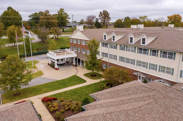 Aerial view of a senior living facility building with a covered entrance, surrounded by green lawns, trees, and pathways. The building has multiple windows and a brown shingled roof. Three flagpoles with flags are visible near the entrance.
