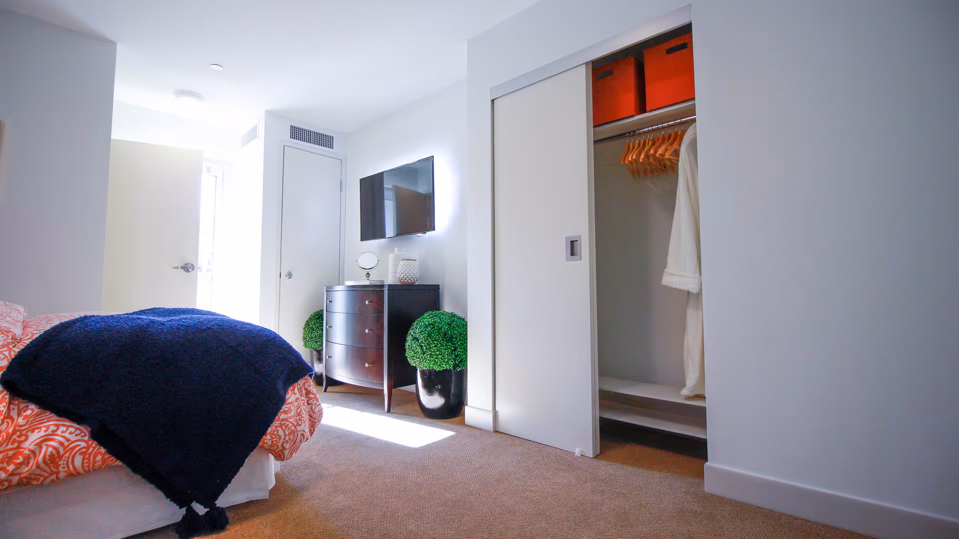 A bright bedroom with a bed covered in orange and white patterned bedding and a dark blue blanket. There is a dark wooden dresser with a round mirror and decorative items on top, a flat-screen TV mounted on the wall above it, and two green potted plants on the floor beside the dresser. The closet has sliding white doors, one of which is open, revealing wooden hangers and a white robe. The room has beige carpet and white walls with a door letting in natural light.