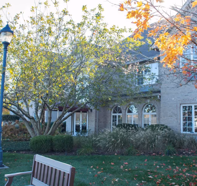 Outdoor view of a senior living facility showing a brick building with arched windows, surrounded by trees with green and orange leaves, a lamppost, and a wooden bench on a grassy area with scattered fallen leaves.