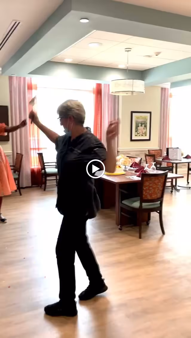 A person wearing a mask dances in a bright dining room with set tables and patterned chairs.
