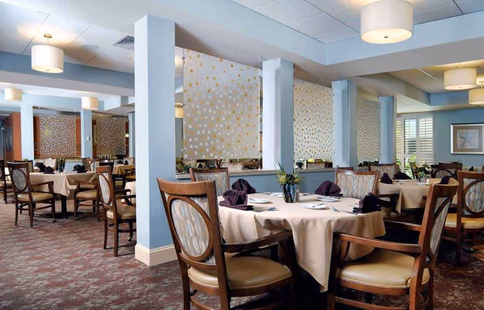 A dining room with round tables covered in beige tablecloths, each set with plates, silverware, and folded dark napkins. Wooden chairs with cushioned seats and backs surround the tables. The room has light blue walls, decorative partitions with a gold and white dot pattern, carpeted floors, and ceiling lights.
