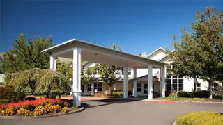 Exterior view of Columbia Place Assisted Living and Memory Care facility showing the main entrance with a covered drop-off area, surrounded by landscaped gardens with colorful flowers and trees under a clear blue sky.