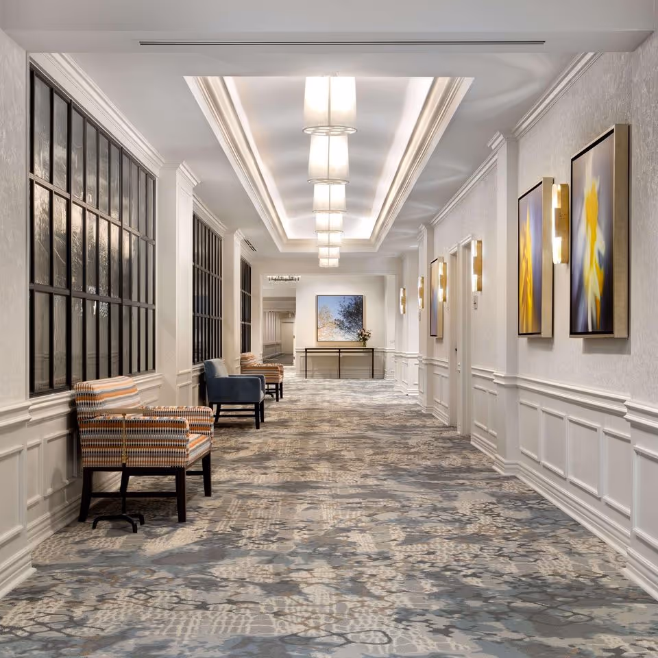 A well-lit, elegant hallway with patterned carpet, white paneled walls, and modern light fixtures on the ceiling and walls. There are several chairs along the left side near large windows, and framed abstract artwork on the right wall. A table with a flower arrangement and a framed picture is visible at the end of the hallway.