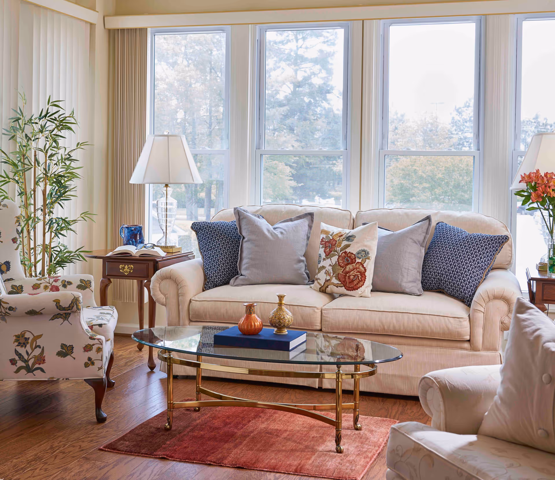 Sunlit living room with a beige sofa, patterned armchairs, a glass coffee table, and large windows overlooking trees.