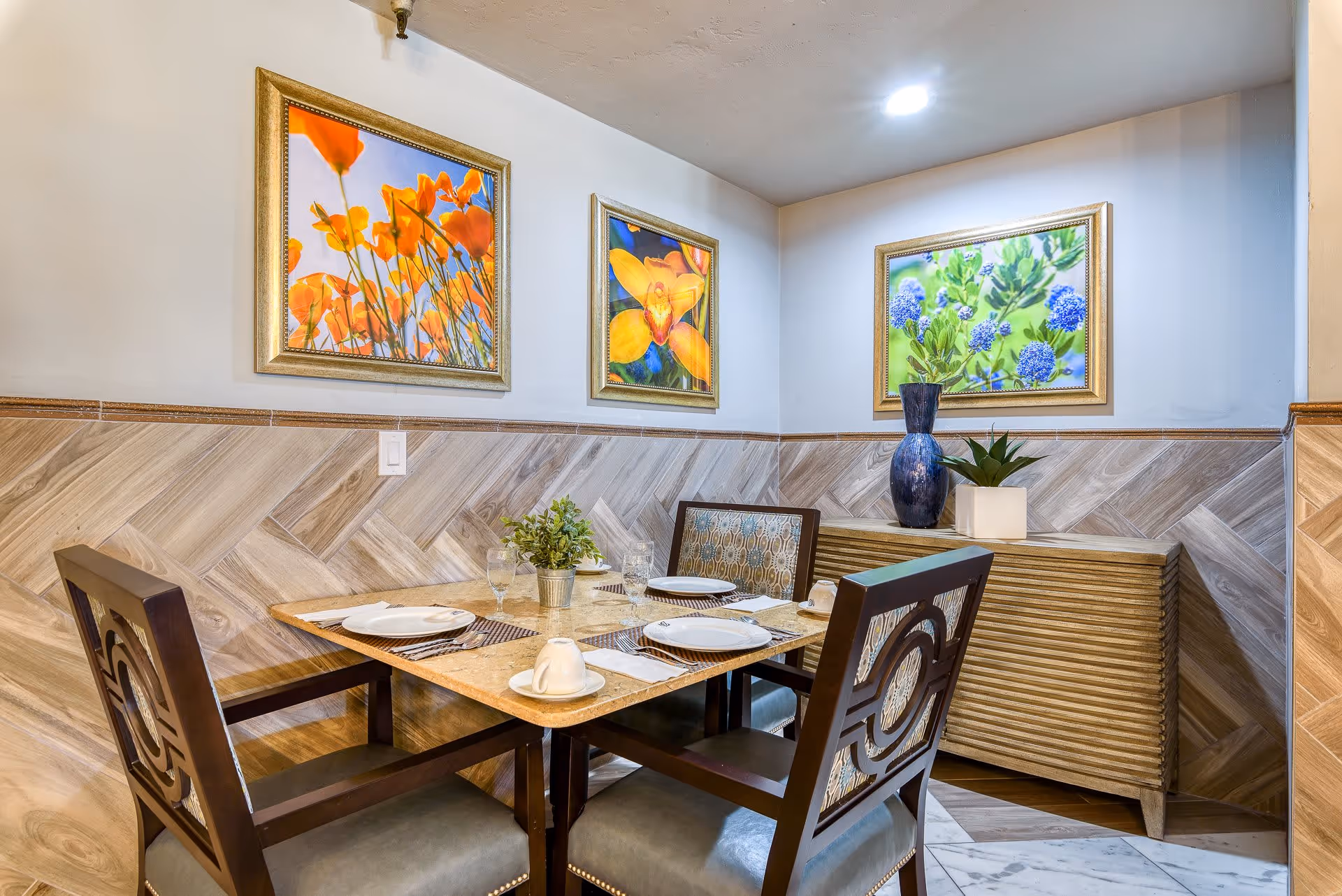 Small dining nook with a table set for four, framed floral paintings on the wall, and a sideboard with decorative vases and plants.