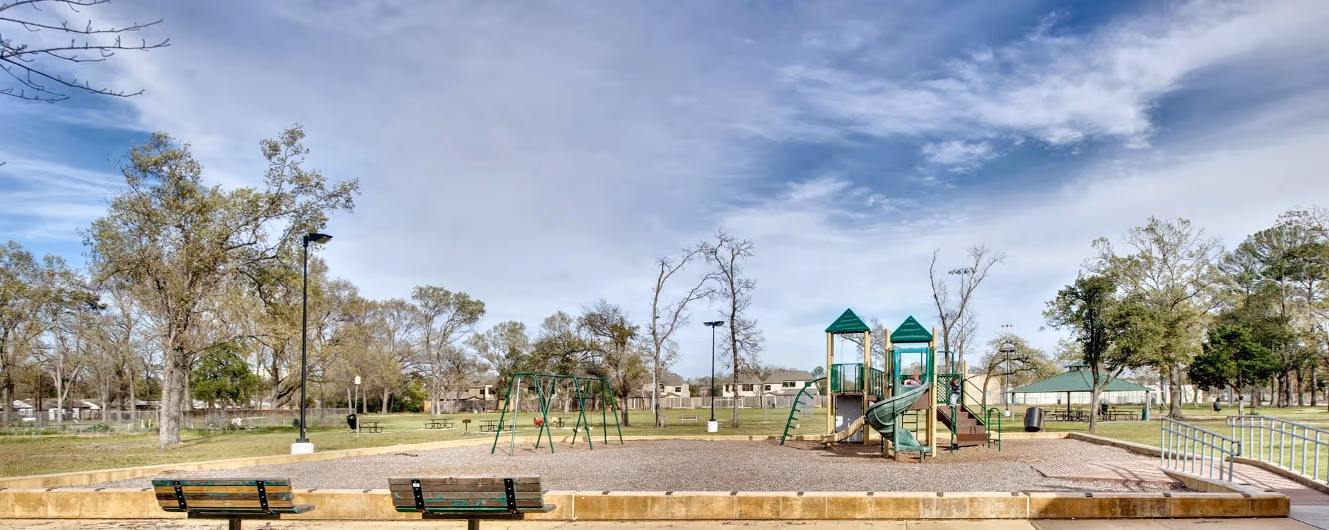 Outdoor playground area with green and beige play structures including slides and swings, surrounded by trees and benches under a partly cloudy sky at Park Shadows Retirement Community.