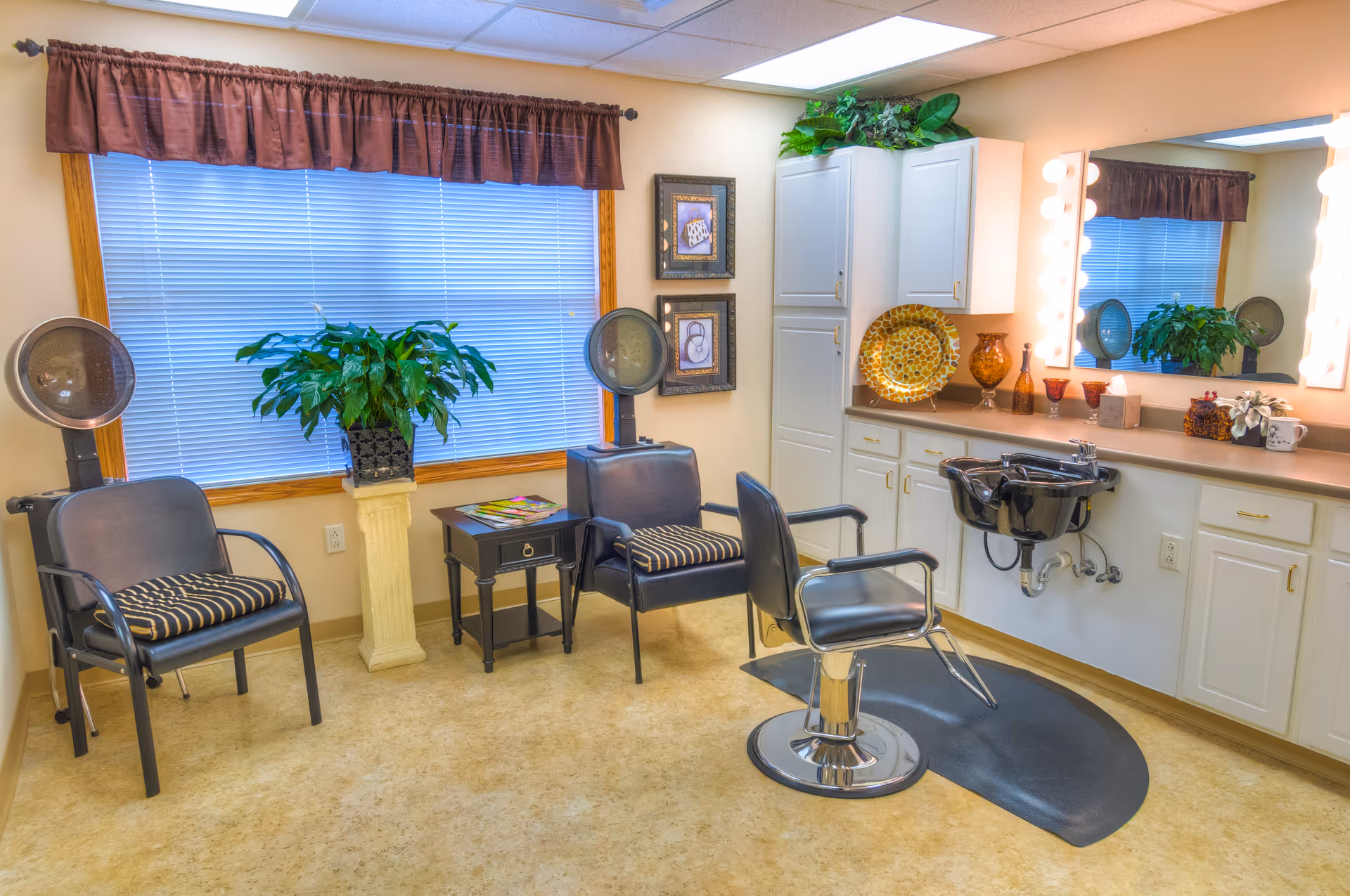 A bright salon-style room with a styling chair and shampoo sink, two hooded hair dryers, waiting chairs, plants, and a mirrored vanity.