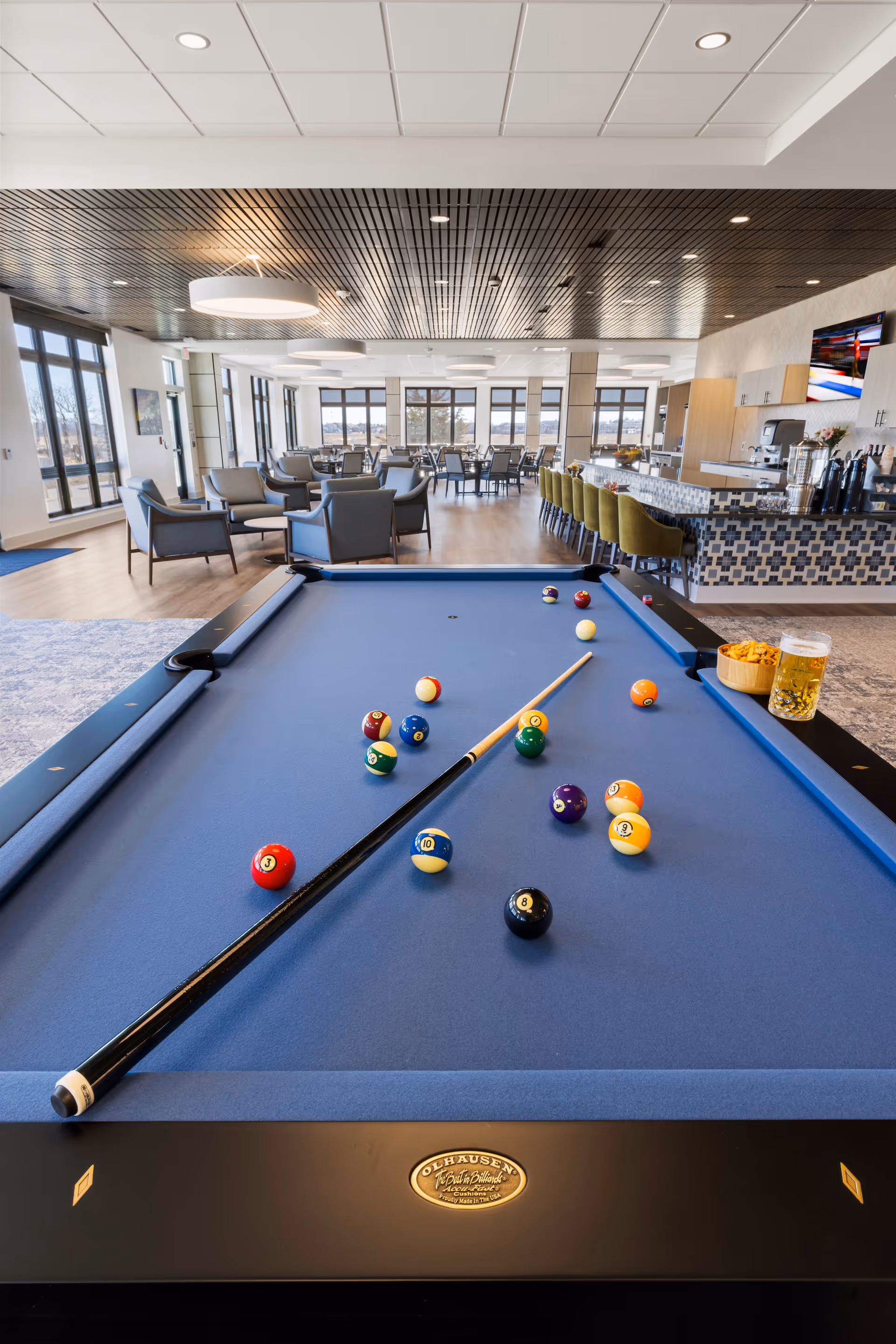 Bright senior living common room showing a blue pool table in the foreground with seating and a bar area in the background.