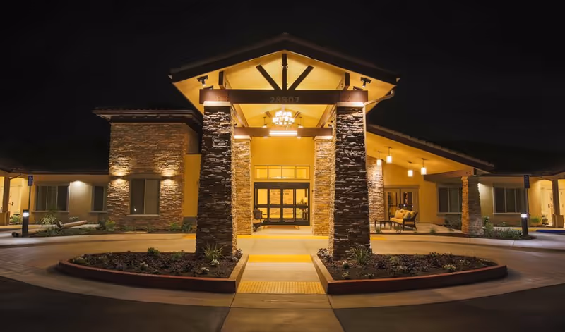Night view of the entrance to a senior living facility with stone pillars, warm lighting, and a covered walkway leading to glass doors. The building has a modern design with a combination of stone and stucco exterior walls.