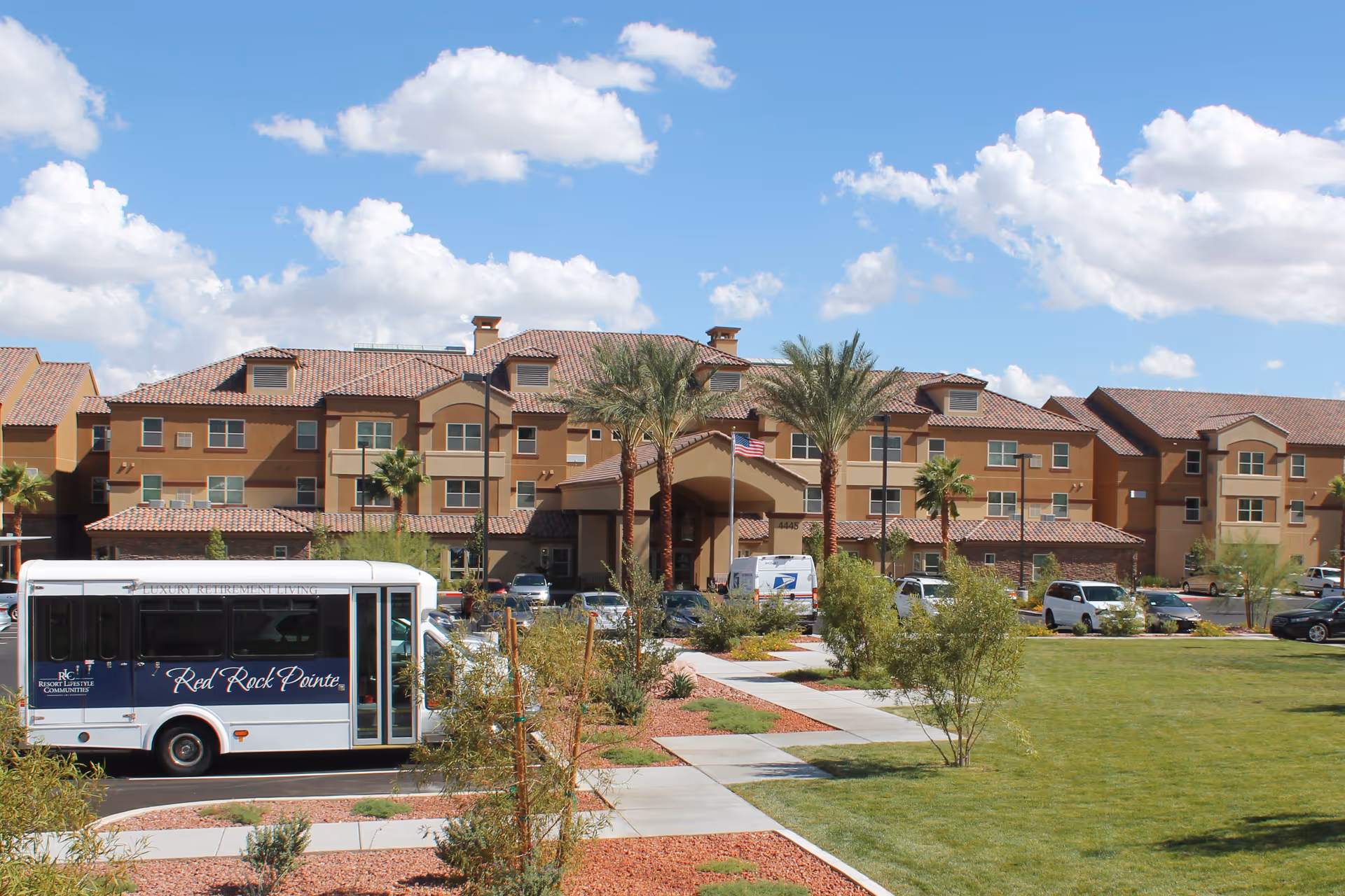 Front view of the Red Rock Pointe multi-story retirement community with a parked shuttle bus, landscaped lawn and palm trees under a blue sky.