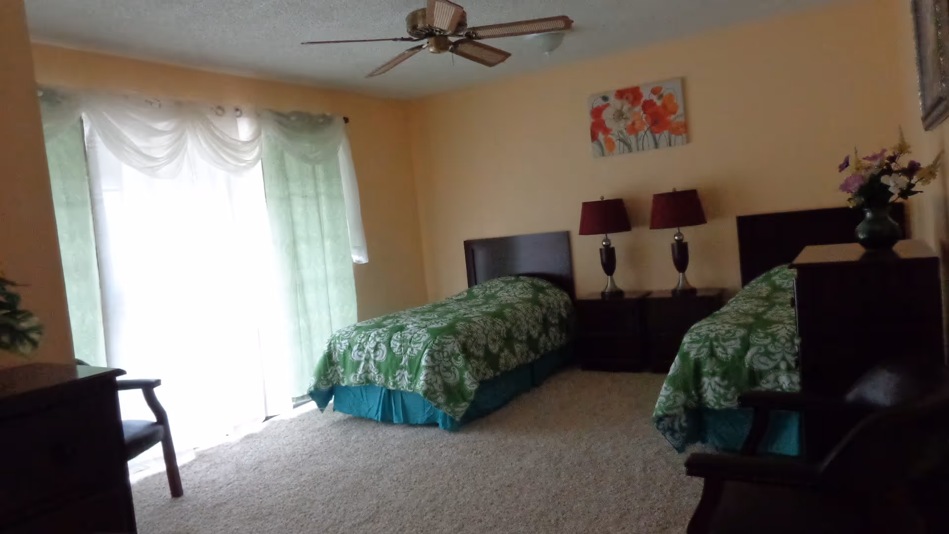 A bedroom with two twin beds covered in green patterned bedspreads. Between the beds is a nightstand with two matching table lamps with red shades. A floral painting hangs on the wall above the nightstand. The room has beige walls, a ceiling fan, and a large window with sheer white and green curtains letting in natural light. There is a chair and a dresser visible in the room.