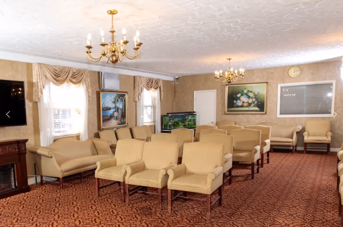 A spacious living room with multiple beige upholstered armchairs and sofas arranged in rows on a patterned carpet. The room features two chandeliers hanging from a textured white ceiling, beige walls with framed paintings, a large flat-screen TV mounted on the wall, and an aquarium. Windows with sheer curtains and valances allow natural light into the room.